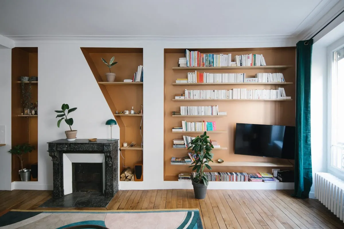 Salon parisien avec bibliothèques encastrées sur mesure au fond ocre, cheminée en marbre noir et téléviseur intégré au meuble