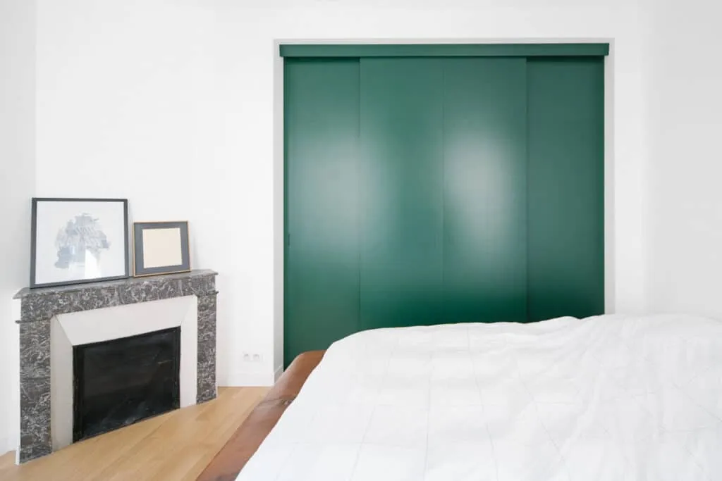Chambre lumineuse aux murs blancs avec grand placard encastré à portes coulissantes vertes, cheminée d’angle en marbre gris, parquet clair et lit au linge blanc