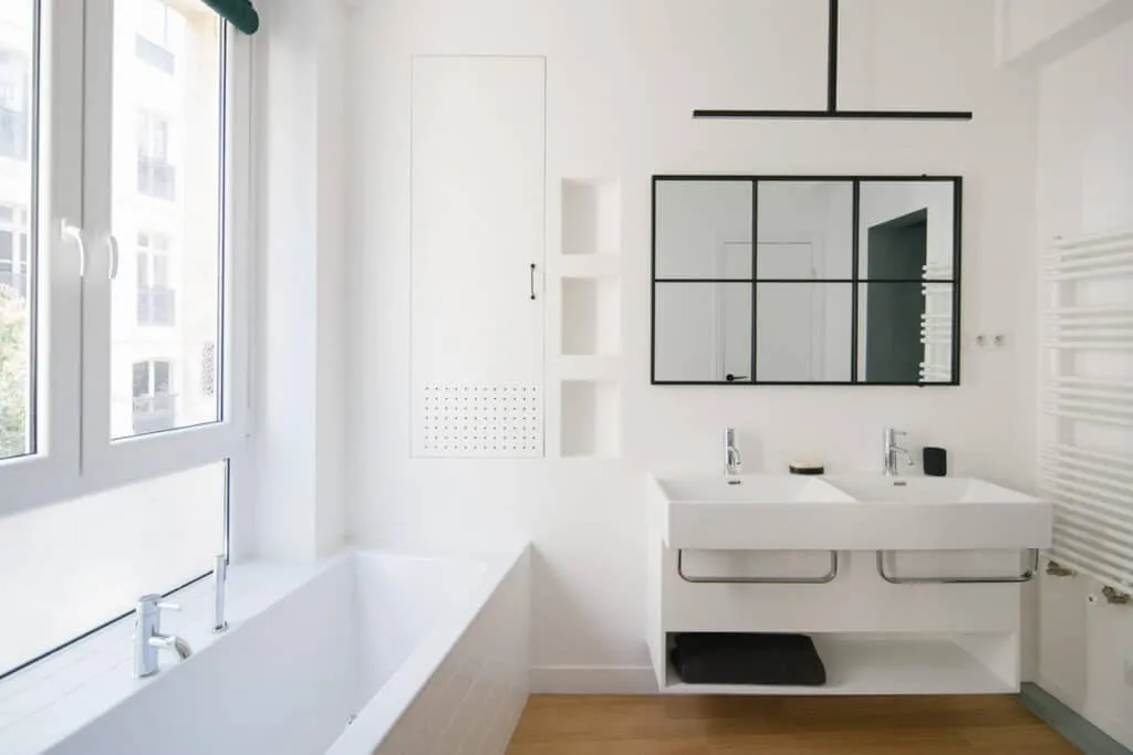 Salle de bain lumineuse aux murs blancs, avec baignoire encastrée sous une grande fenêtre et double vasque suspendue