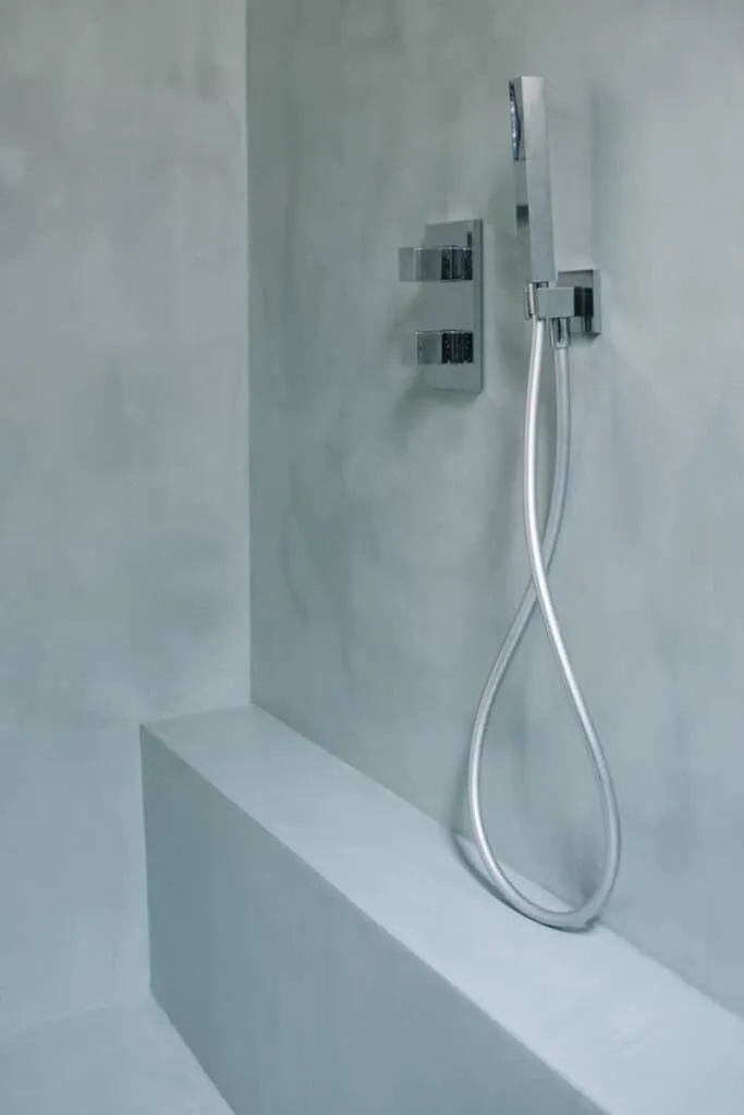 Gros plan sur une douche à l’italienne en béton ciré gris-bleu avec banc intégré, mitigeur thermostatique chromé et douchette à main