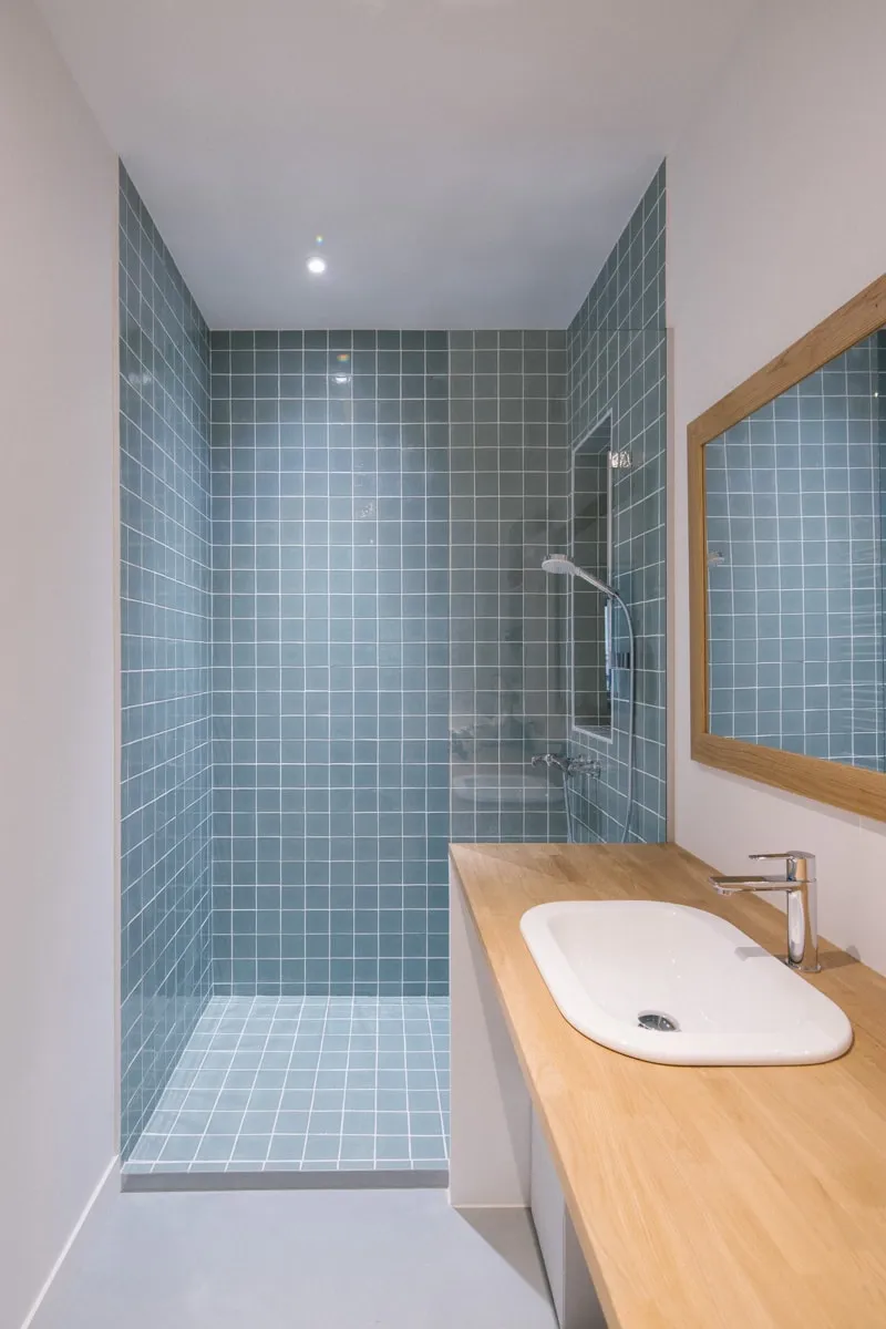 Salle de bain rénovée avec douche à l’italienne en petits carreaux bleu-gris, plan vasque en chêne massif et miroir à cadre en bois