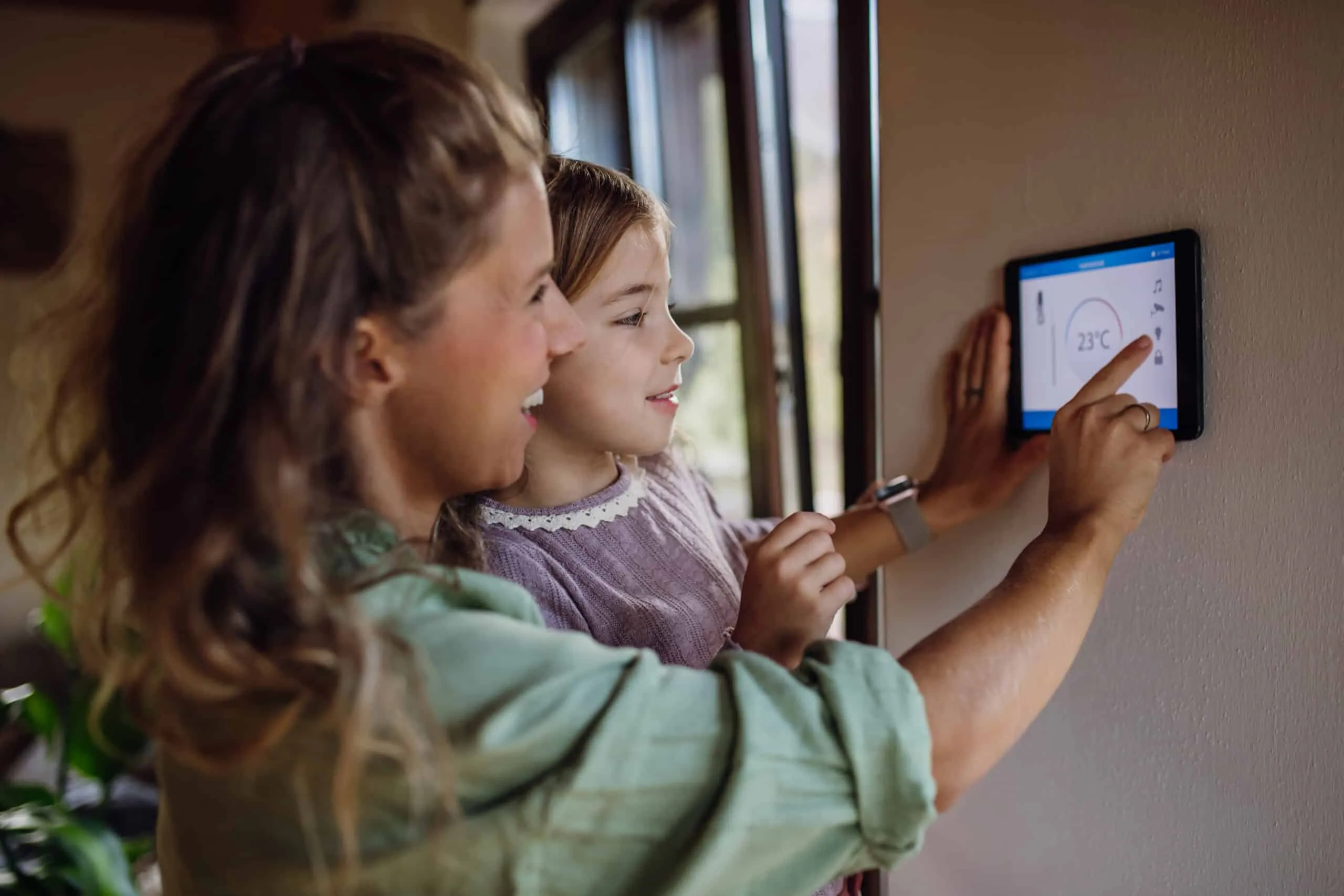 Mère et fille réglant le chauffage à 23 °C sur une tablette murale de domotique