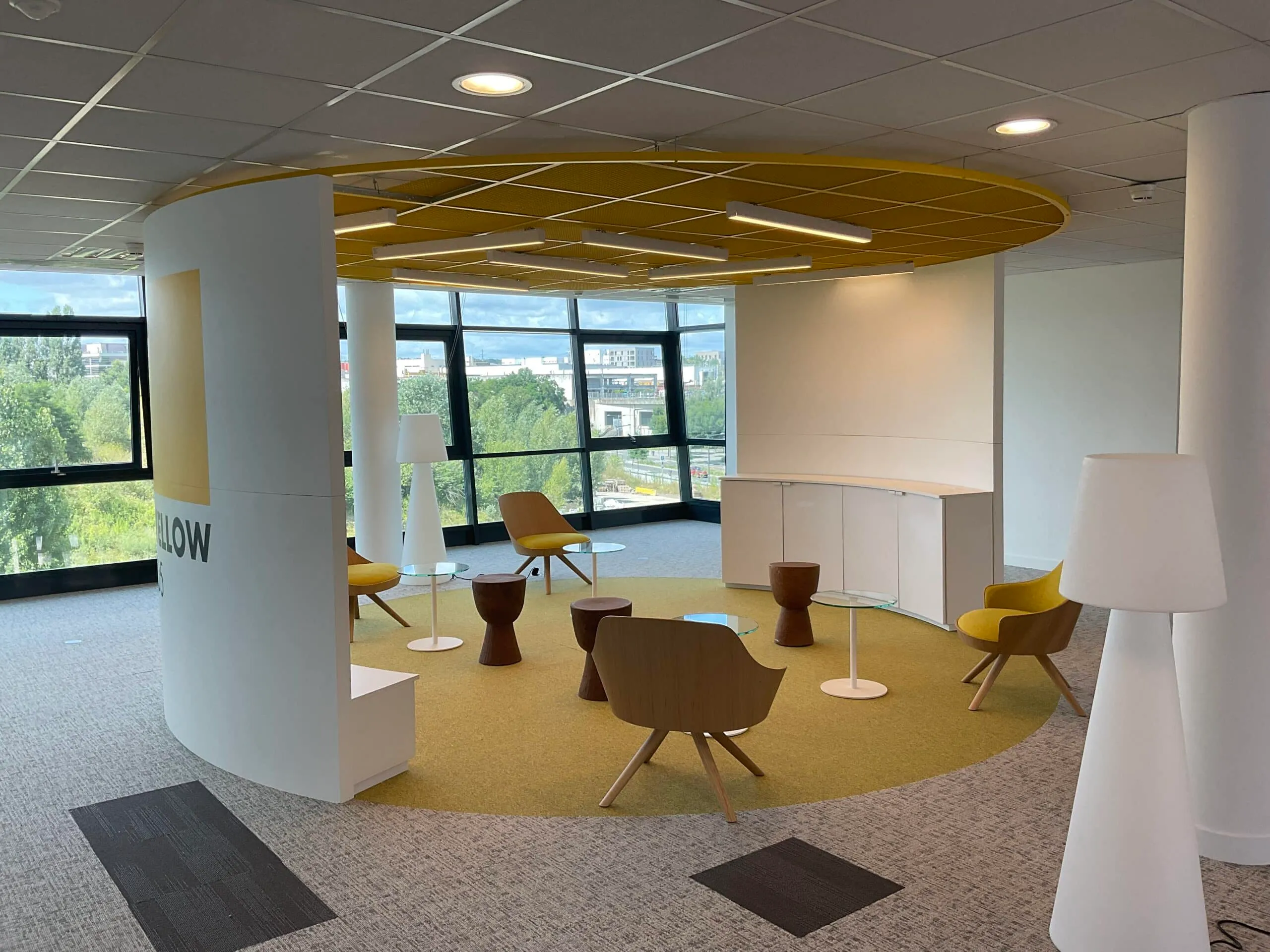 Espace de détente circulaire dans des bureaux aménagés, avec cloison courbe blanche, plafond jaune, fauteuils en bois et moquette jaune