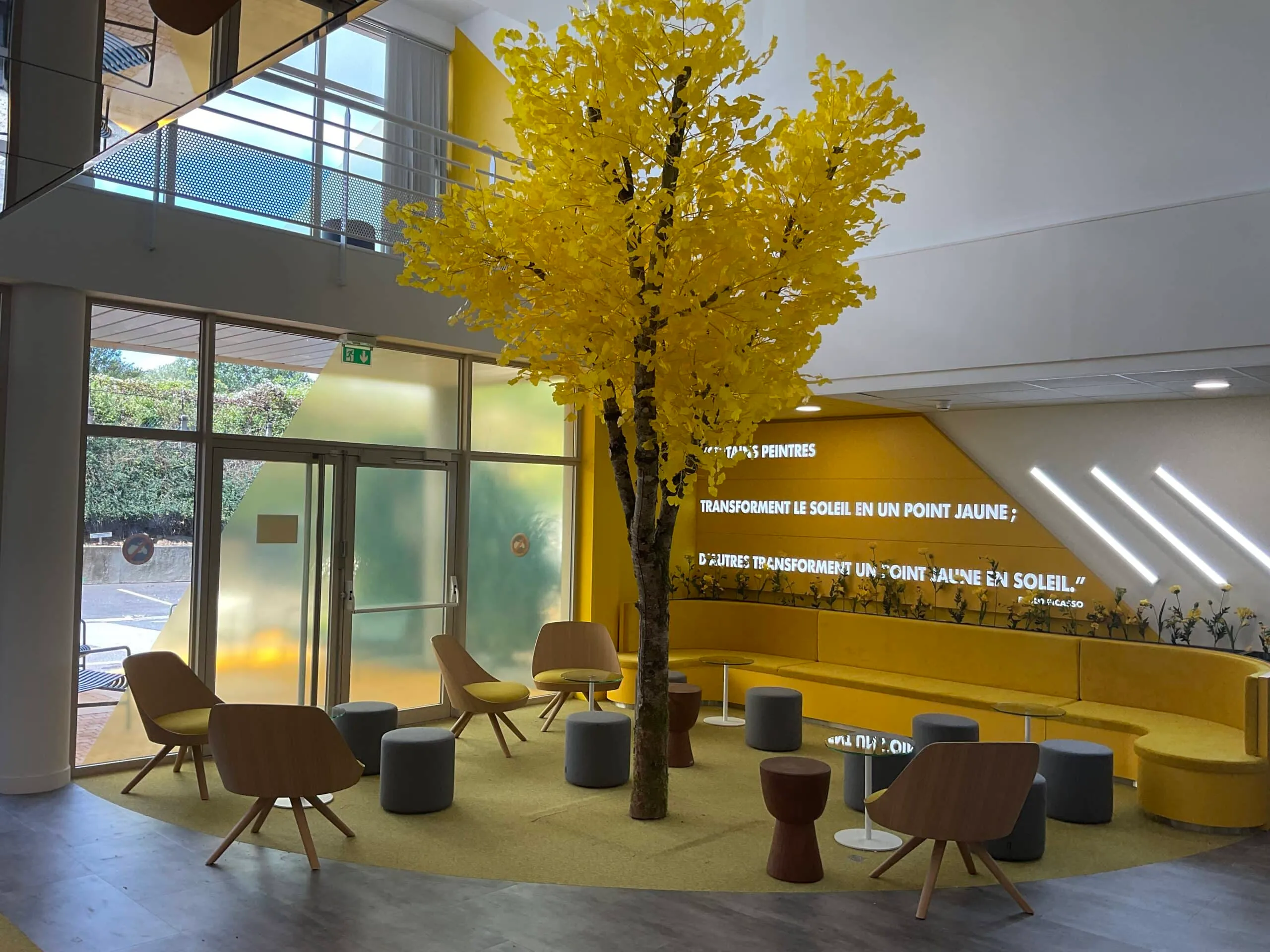 Espace d’accueil aménagé avec un arbre décoratif jaune, une banquette en velours jaune, des fauteuils en bois clair et des pouffs gris