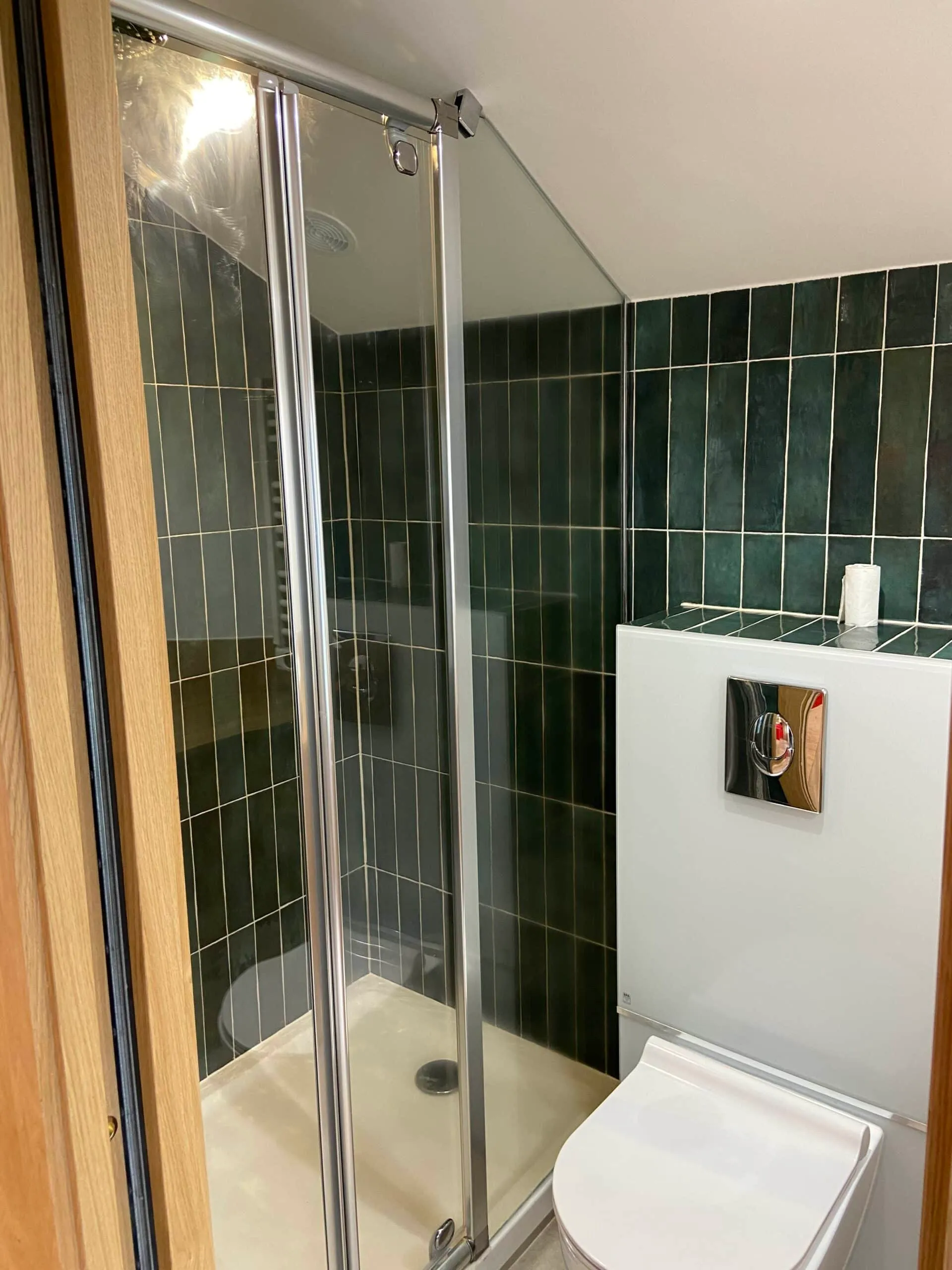 Salle d’eau rénovée sous les combles avec douche vitrée, carrelage vert foncé posé en vertical et WC suspendu blanc