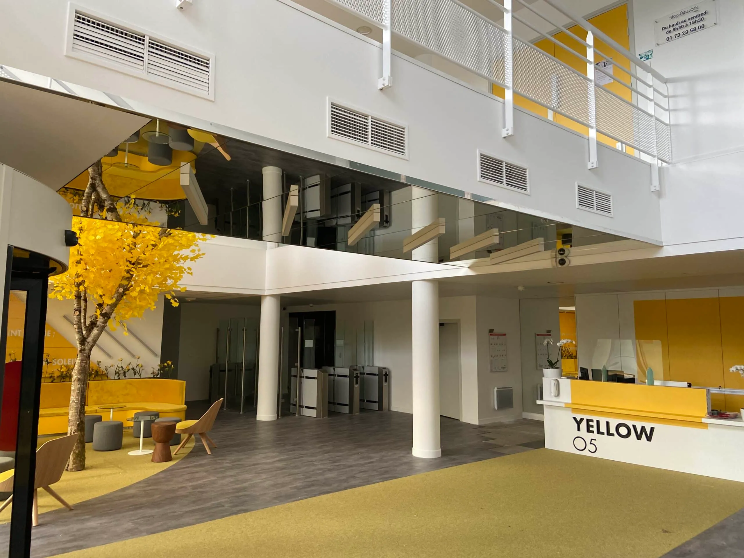 Vaste hall d’accueil d’un immeuble de bureaux avec arbre décoratif jaune, coin salon, comptoir d’accueil blanc et jaune, colonnes blanches