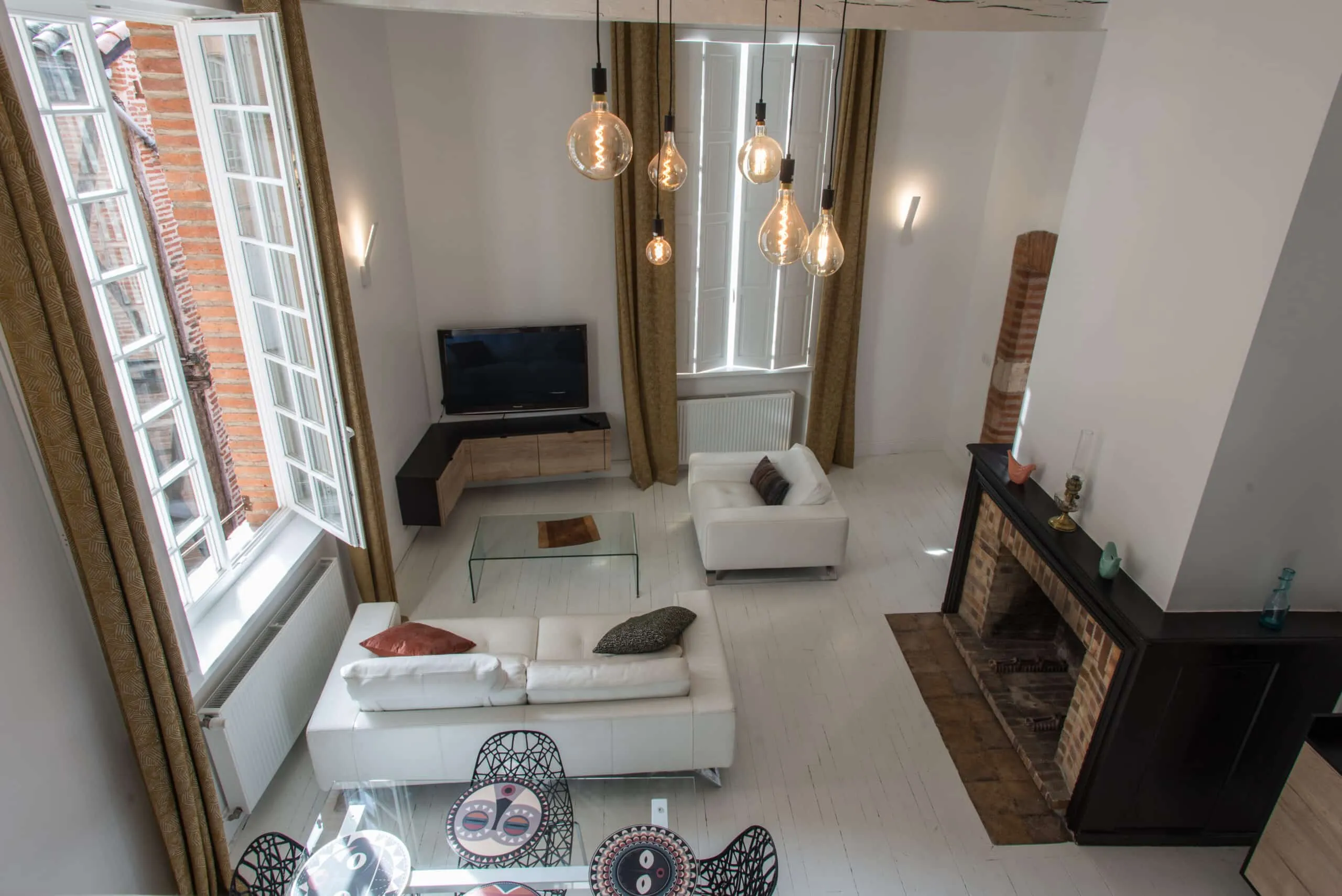 Salon rénové avec parquet blanc, canapés en cuir blanc, cheminée en briques, meuble TV d’angle sur mesure et suspensions à ampoules décoratives