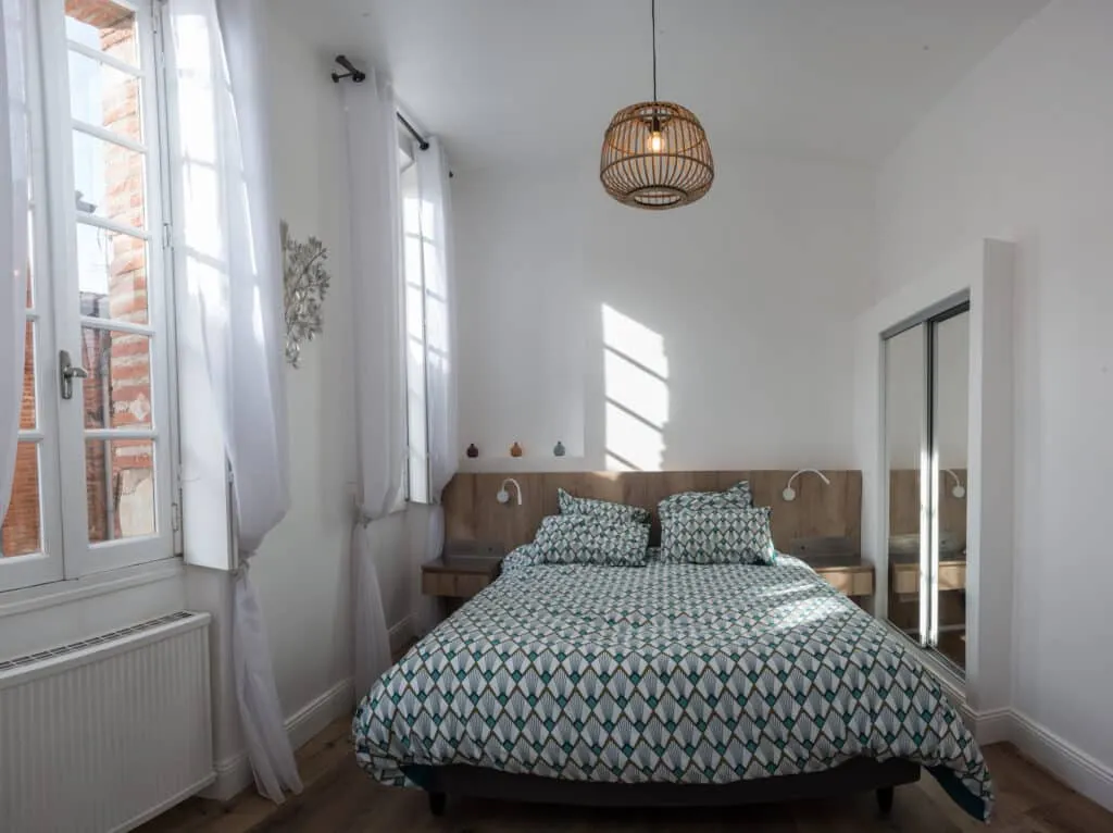 Chambre rénovée aux murs blancs, avec lit double à tête de lit en bois, suspension en rotin et grandes fenêtres à rideaux voilés